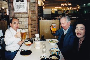 Two older men and a woman sit around a table at a Japanese restaurant in Japan. Charlie and Gordon raise pints of Kirin to the camera, Yumiko smiles.