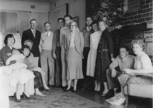 The Parker family and their friends are gathered around a fireplace and pose for the camera. Gordon watches Cherry, who holds baby Kathleen. Harry, Venn, Robin, and June are also in the image.