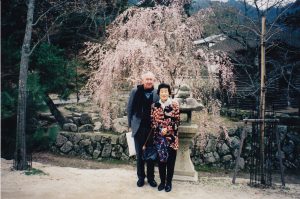 An older couple, Gordon and Cherry Parker, stand in front of a blooming sakura tree.
