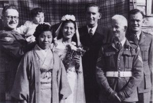Gordon and Cherry Parker, also known as Nobuko Sakuramoto, pose on their wedding day in Japan, with three of their British Commonwealth Occupation Force (BCOF) friends, plus a Japanese woman and their daughter, Margaret. Cherry wears a white wedding dress and holds a posy of flowers, Gordon wears a suit.